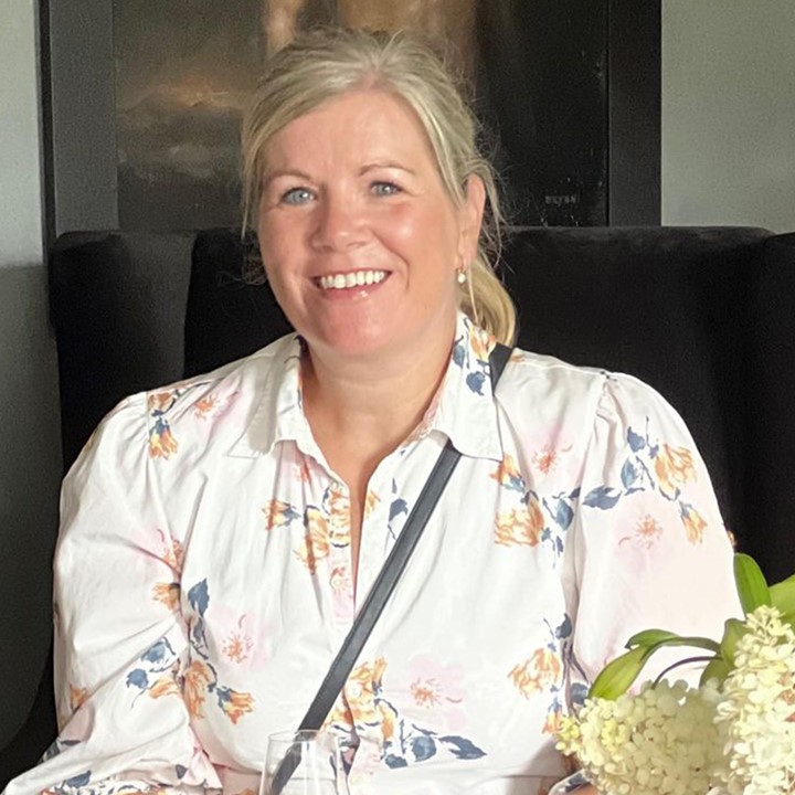 Lise Kjos som har på seg mønstret, lys kjole og har et glass i hånden. Foto.