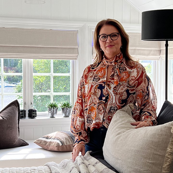 Stylist Berit Vildalen står inne på lyst soverom med oppredd dobbeltseng, vinduer med sjøutsikt og skreddersydde liftgardiner. Foto.