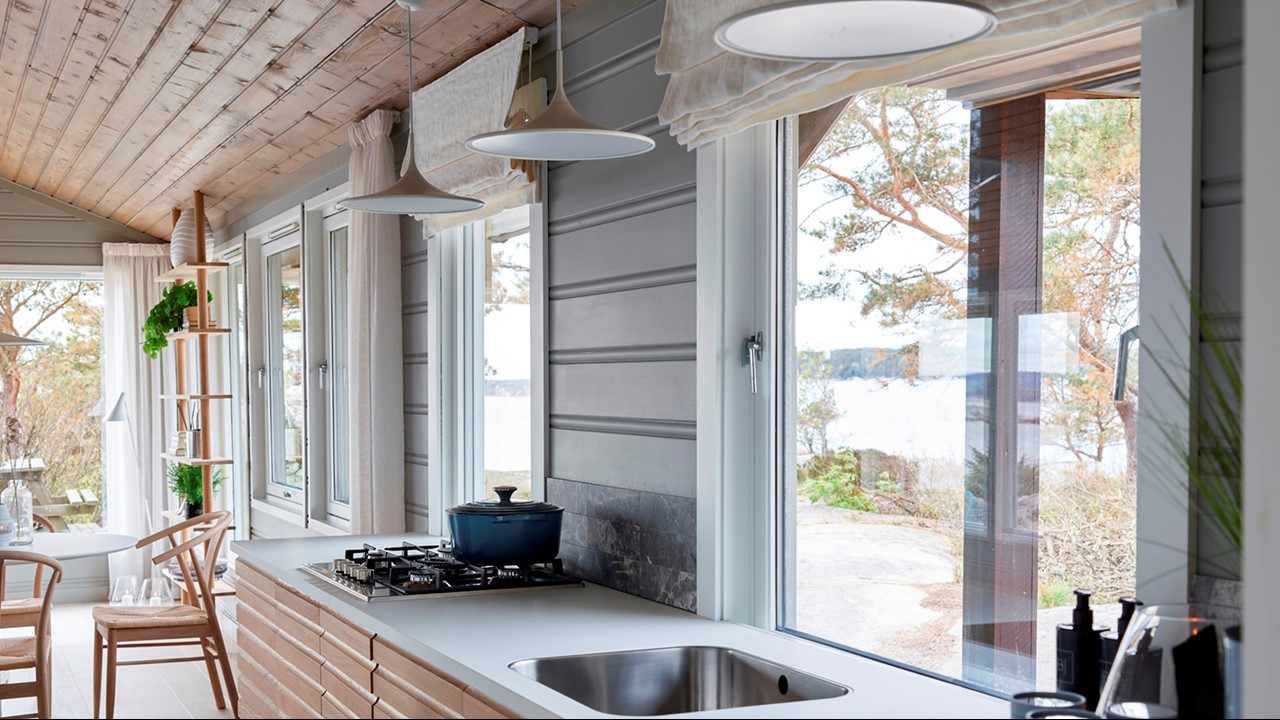 Hytte med grå vegger, kjøkkeninnredning i lyst tre, hvite liftgardiner og gardiner. Fra Eventyrlig Oppussing Fårnøtt. Foto.
