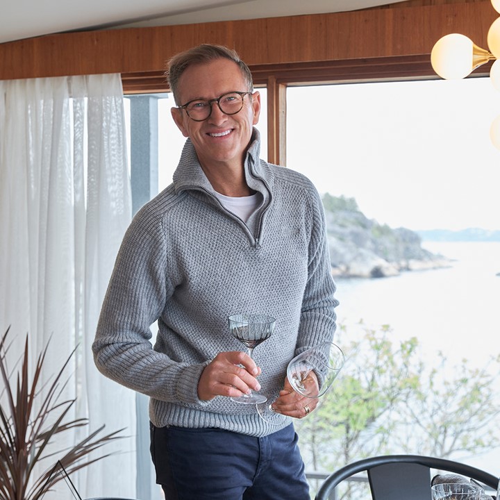 Halvor Bakke står i stuen med store vinduer og gardiner hvite. I hendene holder han to glass. Fra Eventyrlig Oppussing Larvik. Foto.