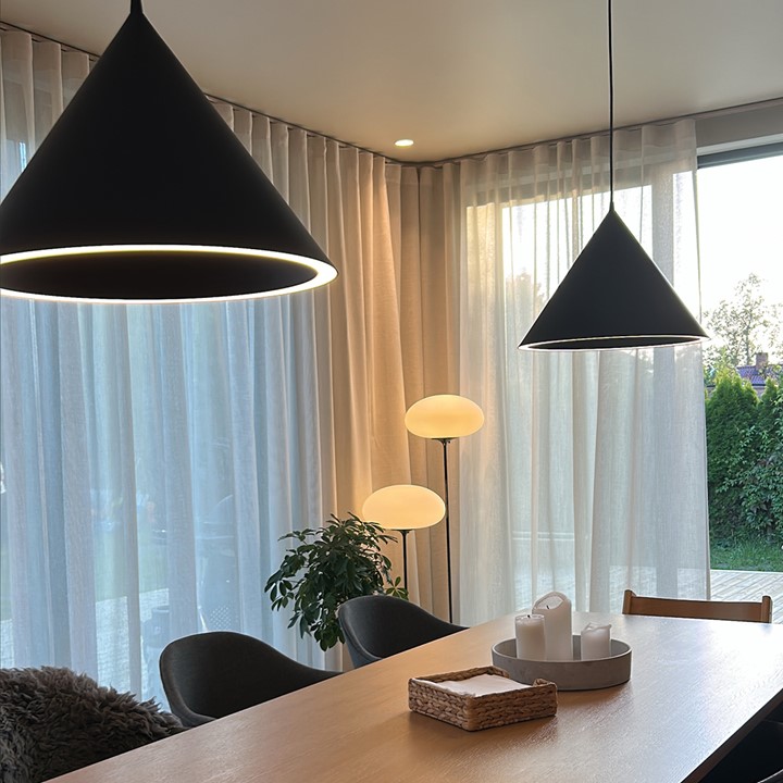Fra spisestue ser vi store vinduer med florlette gardiner i taket, spisebord med stoler, to kjegleformede taklamper og to stålamper. Foto.