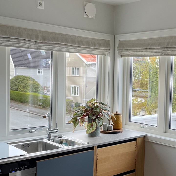 Lyst kjøkken med store vinduer med skreddersydde, lyse liftgardiner montert innvendig i karm, kjøkkeninnredning med fronter i tre og blått. Foto.
